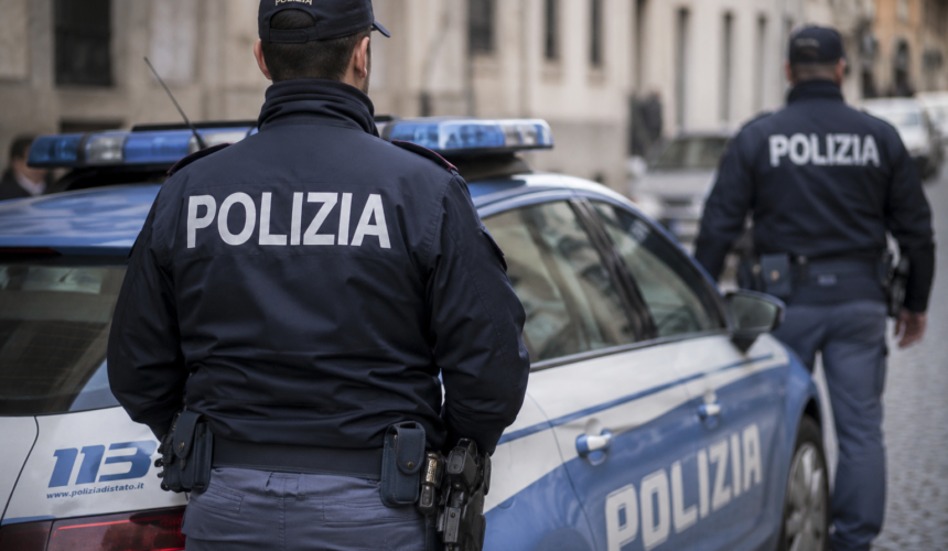 Due poliziotti italiani in uniforme accanto a un’auto della Polizia di Stato parcheggiata in una strada cittadina.