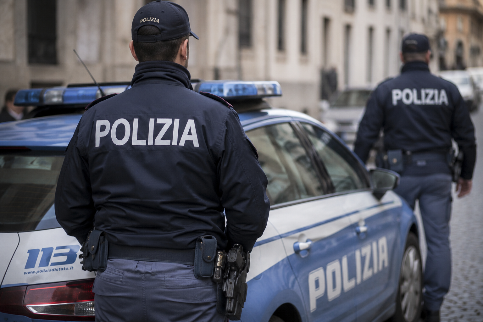 Due poliziotti italiani in uniforme accanto a un’auto della Polizia di Stato parcheggiata in una strada cittadina.