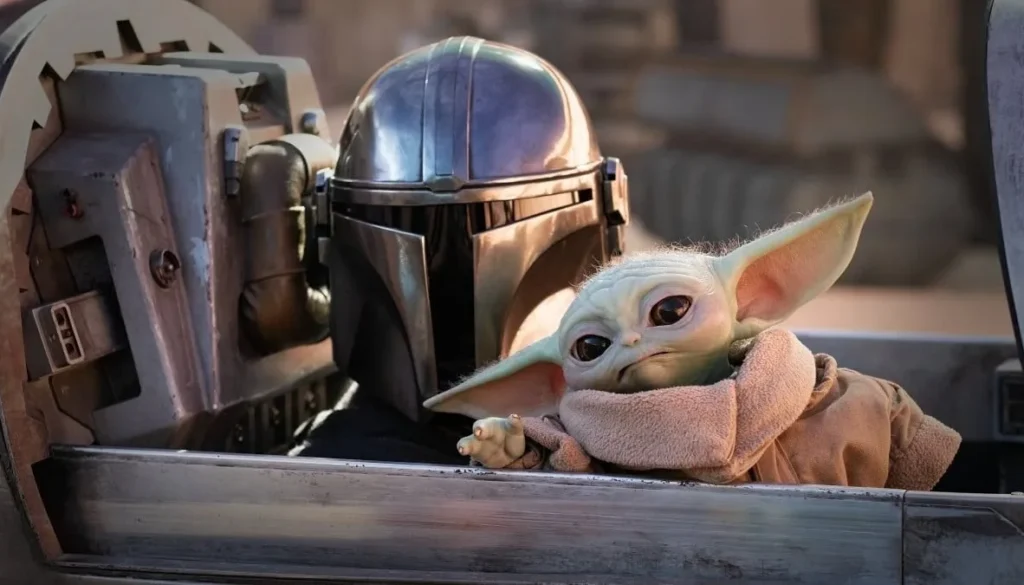 Il Mandaloriano con armatura argentata accanto a Grogu, detto Baby Yoda, seduti a bordo di un veicolo nell’universo di Star Wars.