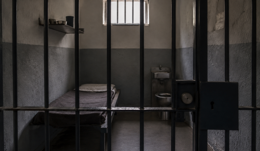 Cella di carcere con letto singolo, lavabo e sbarre metalliche in primo piano, illuminata da una piccola finestra con grate.