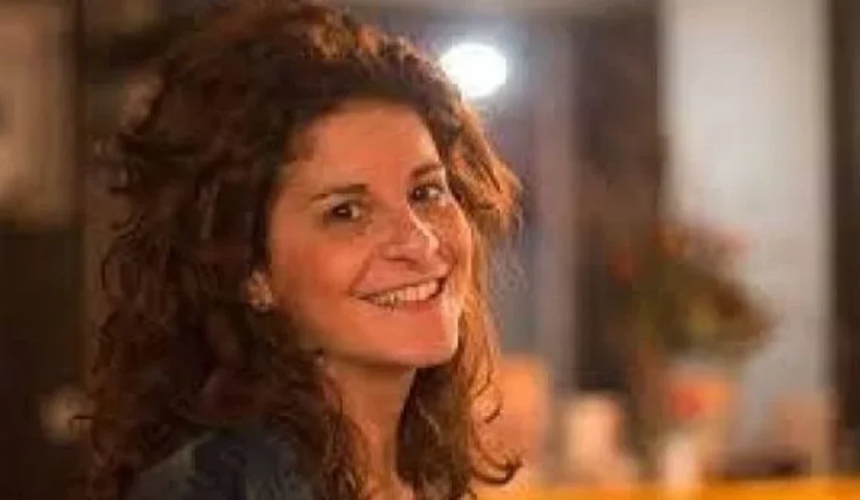Donna con capelli ricci castani che sorride verso la fotocamera in una stanza illuminata con tonalità calde.
