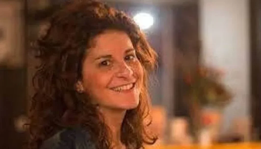 Donna con capelli ricci castani che sorride verso la fotocamera in una stanza illuminata con tonalità calde.