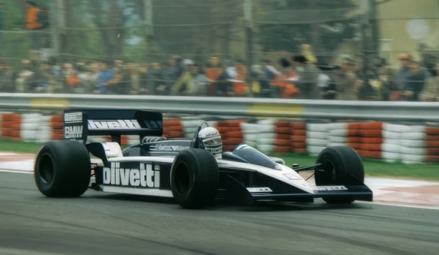 Auto storica di Formula 1 con livrea nera e bianca percorre una curva del circuito davanti agli spettatori dietro le barriere.