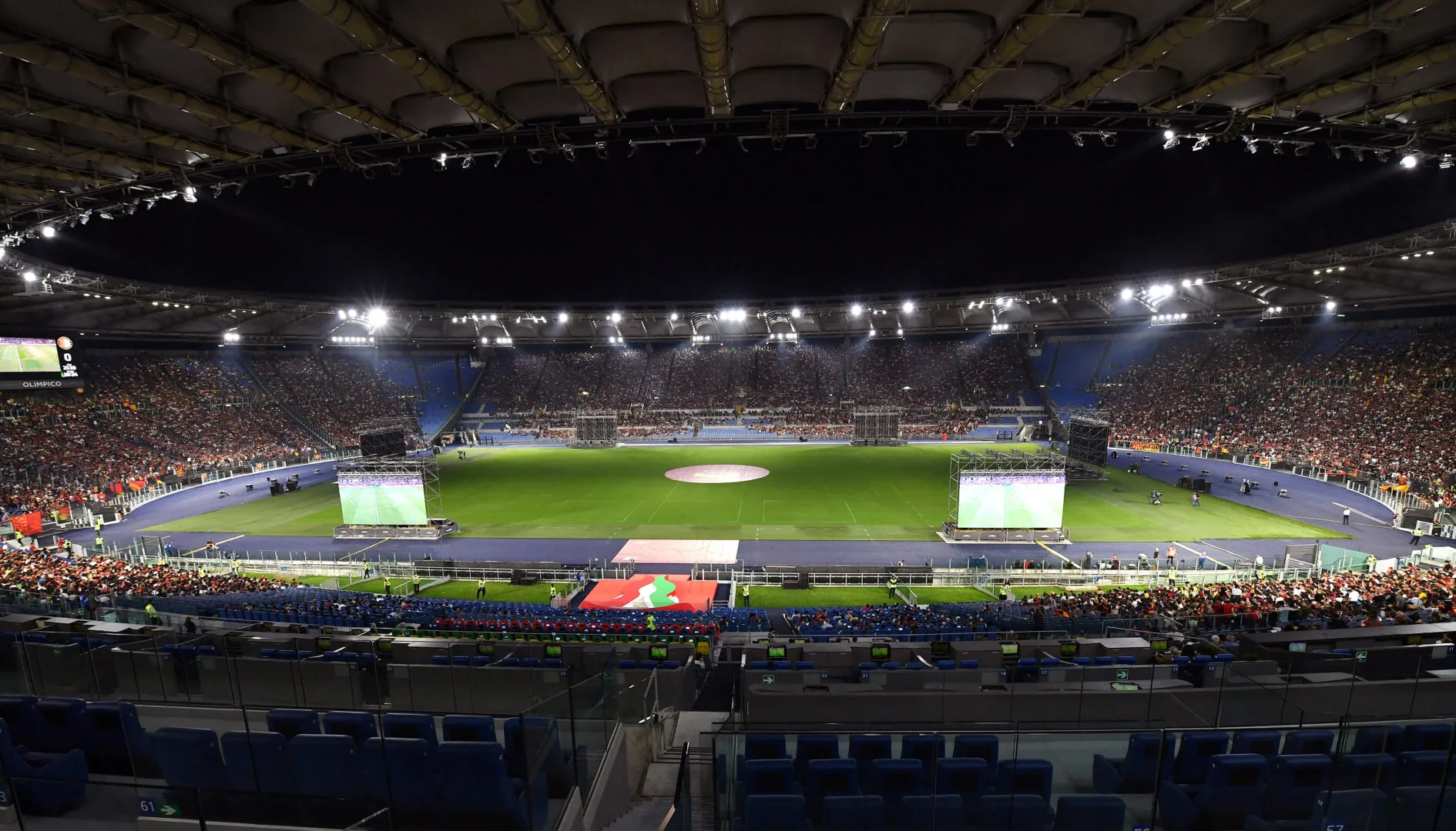 Vista panoramica dello Stadio Olimpico pieno di pubblico durante un evento sportivo serale con il campo illuminato dai riflettori.