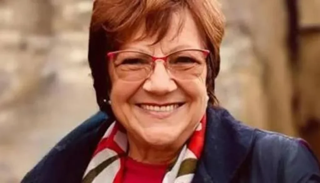 Donna con capelli corti castani e occhiali rossi che sorride verso la fotocamera in un ritratto ravvicinato.