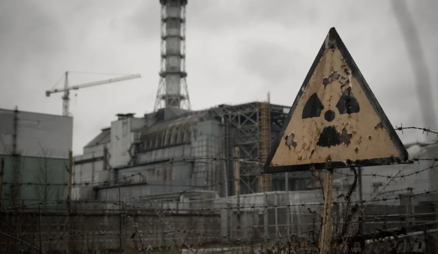 Cartello di pericolo radioattivo arrugginito davanti a una centrale nucleare circondata da recinzioni e filo spinato sotto un cielo grigio.
