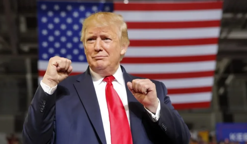 Donald Trump in abito scuro e cravatta rossa con i pugni alzati davanti a una grande bandiera americana.