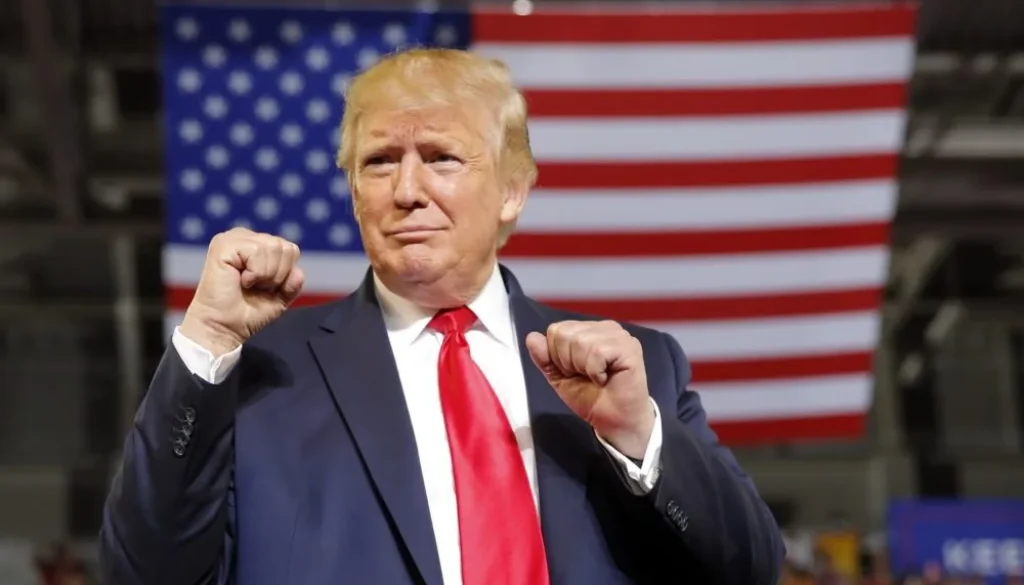 Donald Trump in abito scuro e cravatta rossa con i pugni alzati davanti a una grande bandiera americana.