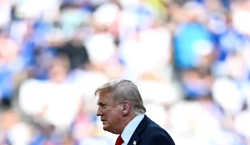 Donald Trump in abito scuro e cravatta rossa ripreso di profilo durante un evento pubblico con il pubblico sfocato sullo sfondo.