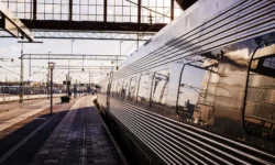Treno moderno fermo in una grande stazione ferroviaria illuminata dalla luce calda del tramonto, con riflessi metallici sulle carrozze e binari che si estendono in prospettiva.