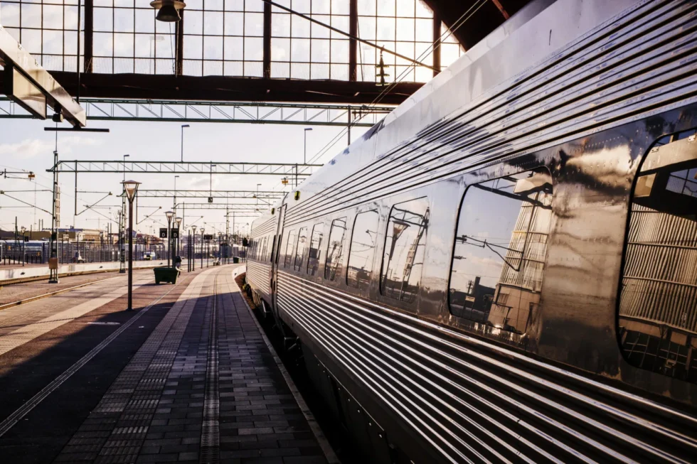 Treno moderno fermo in una grande stazione ferroviaria illuminata dalla luce calda del tramonto, con riflessi metallici sulle carrozze e binari che si estendono in prospettiva.