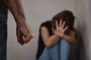 Donna seduta a terra in atteggiamento di difesa mentre al centro dell'immagine si vede in primo piano il pugno chiuso di un uomo, simbolo della violenza di genere e della paura vissuta dalle vittime.