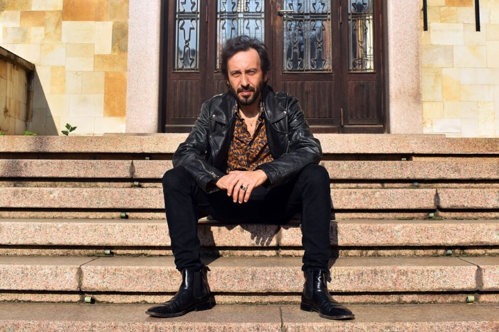 Uomo seduto sui gradini di un edificio con giacca in pelle nera e camicia fantasia, ritratto in un contesto urbano.