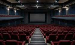 Interno di una sala cinematografica vuota con file di poltrone rosse rivolte verso uno schermo spento e una balconata superiore.