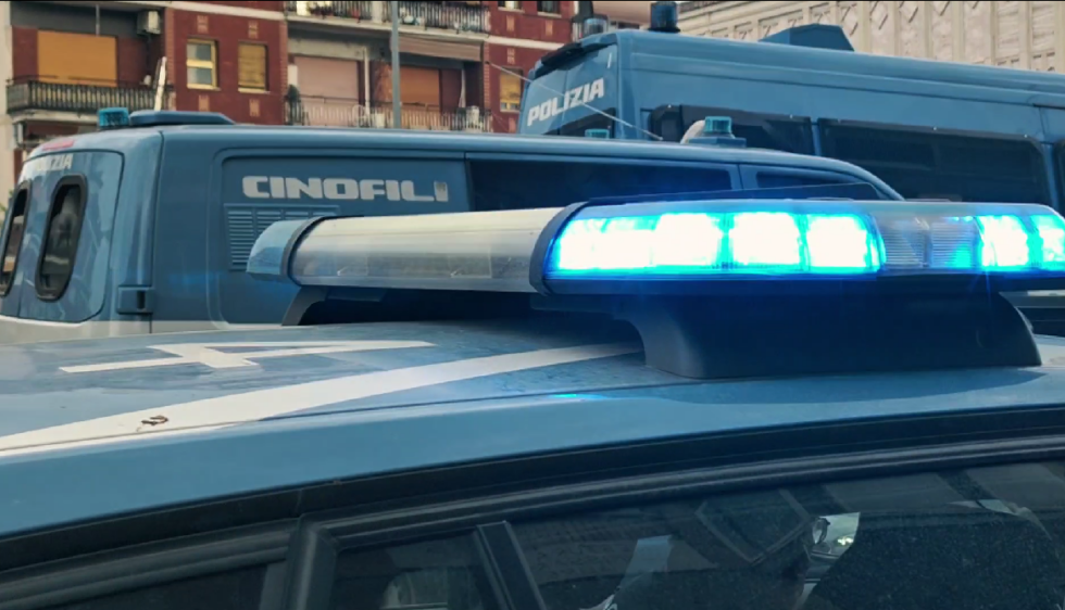 Primo piano del lampeggiante blu di un’auto della Polizia di Stato, con furgoni della polizia sullo sfondo.