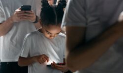 Bambina con chignon concentrata a guardare lo schermo illuminato del cellulare circondata da persone.