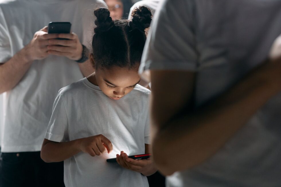 Bambina con chignon concentrata a guardare lo schermo illuminato del cellulare circondata da persone.
