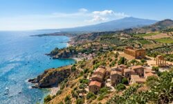 Vista aerea di un paesaggio costiero con mare blu, templi dorici in stile greco antico su una collina, borgo con case in pietra e una montagna sullo sfondo.