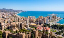 Vista panoramica dall'alto di una città costiera con mura fortificate in primo piano, una cattedrale, un'arena circolare, edifici urbani e il mare sullo sfondo.