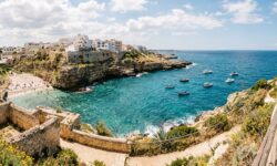 Baia marina rocciosa con spiaggia sabbiosa, edifici bianchi arrocati su una scogliera e barche a remi sul mare turchese